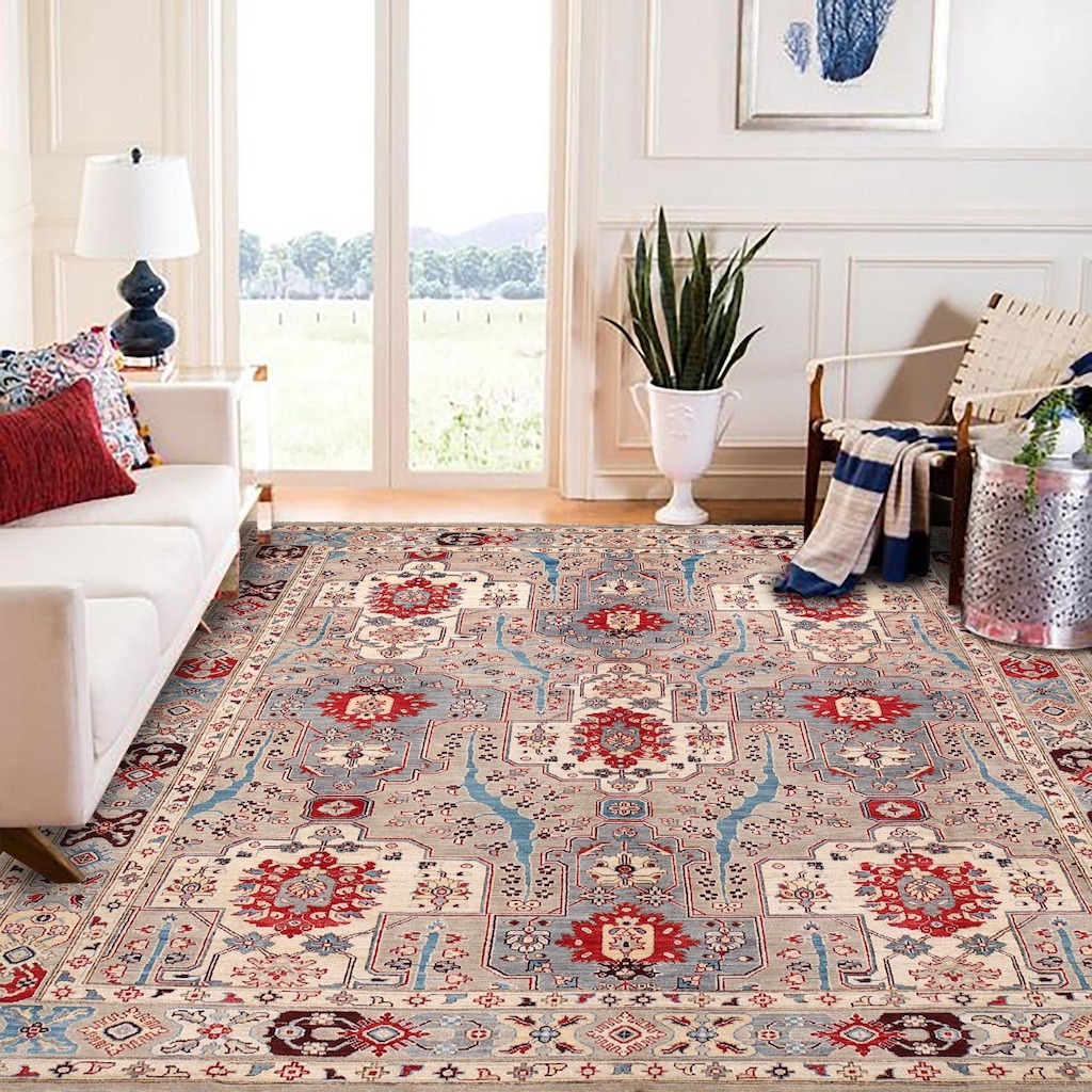 Bright, airy living room with a large patterned area rug in red, blue, and cream tones. A white sofa with colorful pillows sits on the left beside a lamp, while a wooden chair with a draped blanket and a potted plant are on the right. Tall glass doors in the background open to a scenic outdoor view.